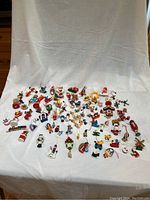 Wide view of more than 70 small, colorful wooden Christmas ornaments scattered on white cloth background.
