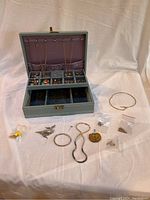 Open jewelry box with necklaces hanging inside and various jewelry pieces arranged outside including bracelets, brooch, and earrings in plastic bags.