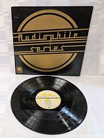 Photo of vinyl record with visible grooves and center label, laid flat next to its cover labeled 'Audiophile Series' in black and gold design.