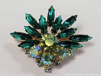 Front view of MCM brooch pin showing emerald green marquise rhinestones arranged in burst pattern with large central faceted aqua green rhinestone and smaller iridescent rhinestones below.