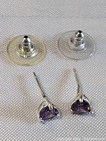 Pair of sterling silver stud earrings with triangular amethyst stones, shown with clear plastic backing discs on a textured fabric background.