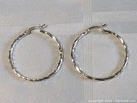 Pair of sterling silver carved hoop earrings laid flat on a white textured surface showing size and clasp detail.