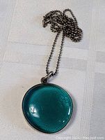 Sterling silver ball chain with round blue-green domed art glass pendant on white textured surface, close-up.