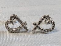 Pair of sterling silver stylized heart shaped stud earrings with clear crystals on front surface, shown from front under good lighting on white textured background.