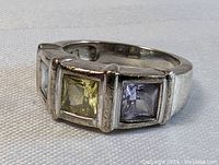 Three-quarter side view of a sterling silver ring with three square-cut stones showing light blue topaz, yellow-green tourmaline, and light purple amethyst.