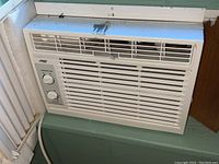 Front view of white Arctic King window air conditioner installed in window frame showing vents and control panel.