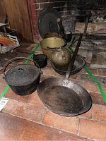 Photo showing collection of various antique cast iron and metal pots, frying pan, small bucket, and stoneware jug placed on a tiled floor next to a fireplace.