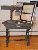 Photo of vintage wooden side table, banker's lamp on table, one framed print leaning against wall on table