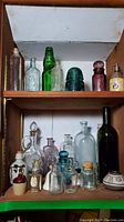 Shelf with multiple vintage glass bottles of various colors and sizes including green insulator and purple mini bottle alongside pottery and bells.