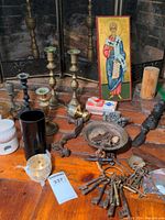 Photo shows four brass candlesticks, religious icon, black vase, rings of skeleton keys, and small assorted figurines on a wooden surface by brick hearth.