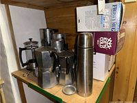 Shelf with multiple stove top espresso makers, thermal bottle, and boxed insulated mugs