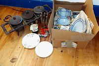 Wide view showing ceramic pots on metal rack, knife block with cutlery, two white plates, and a cardboard box with ceramic dishes.