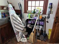 Photo showing ironing board with cover, white iron, broom, mop handle, shelving units with cleaning supplies and tools in corner of room.