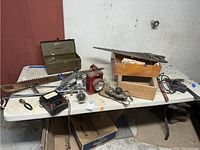 Wide view of table with large green metal toolbox open, vintage hand tools including saw blade, flashlight, wooden toolbox, plugs, and battery charger.