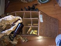 Top view of a cardboard divider box containing vintage amber bottles, a small ceramic jar, and crumpled packing paper. Additional vintage bottles and items can be seen in the background on the wooden floor.