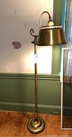 Full view of vintage brass toned metal floor lamp with curved arm and round base, standing next to a green wainscoted wall