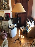 Full view of the vintage floor lamp standing on wooden floor among other household items, showing metal tripod base and beige fabric shade