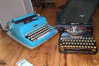 Two vintage manual typewriters side by side on wooden floor, one blue Royal in plastic box and one black Corona Four with metal box.