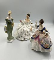 Three Royal Doulton lady figurines shown together: one in green dress, one in floral dress, one in white dress with gold accents.