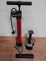 Red upright Wrench Force hand air pump with pressure gauge, hose, and large stable base, next to compact black foot pump and black tubular pump.