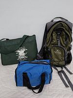 Photo showing three bags: a green Teton Sports backpack, a green NRA shoulder bag with white logo, and a small blue insulated zippered bag, all used condition.