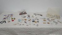Overview of all jewelry making supplies including key rings, earring hooks, chains, loose beads in plastic compartments and bags, and assorted small components laid out on white quilted surface.