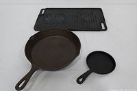 Three cast iron items shown: medium frying pan, small fry pan and rectangular grilling skillet with ribbed surface and handles.