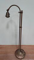 Full view of the copper swan neck floor lamp standing on a wooden floor against a white wall. Shows the lamp's overall structure including base, support column, swan neck arm, and light fixture.