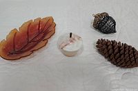 Four fall-themed decor items: amber glass leaf dish with visible veining, alabaster trinket dish with acorn-shaped lid, metal acorn ornament, and natural pine cone on white surface.