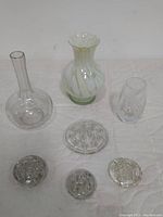 Several glass items including one cream and white swirl vase, a clear glass decanter with long neck, a clear cut crystal vase with etched fern leaf pattern, and four glass flower frogs (one large, two medium, and one small) arranged on a white surface.