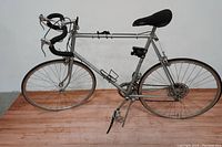 Side view of the full silver Fuji Monterey men's road bike on wooden floor against white wall showing frame, tires, drop handlebars, saddle and pedals.