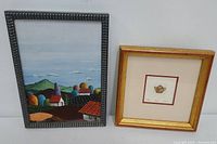 Overview of the two framed artworks side by side on a white surface, the Salvadorian landscape painting on the left and the teapot etching on the right.