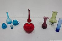Front view of the nine colored glass items arranged on white surface, showing various vase shapes and colors including light blue, red, amethyst, avocado green, and cobalt blue pieces.