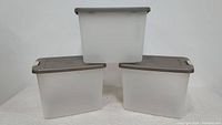 Three Sterilite translucent plastic shelf tote containers with gray lids arranged in a stacked formation against a white backdrop.