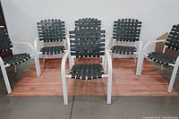 Set of six outdoor chairs with white metal frames and dark green vinyl webbing. Chairs arranged on wooden flooring against plain white background.