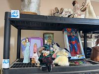Photo of shelf with various dolls including Superman doll in box, SpongeBob Barbie box, plush clown, and other dolls