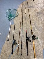 Four fishing rods laid out on ground with a fishing net. Rods are various types and sizes with attached reels. Net has white handle and green netting.