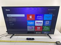 42-inch Hisense Roku TV screen showing Roku home menu with multiple streaming apps visible. TV is switched on and on a white surface with remote control in front.