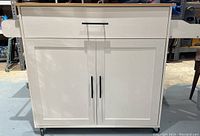 Front view of the white rolling kitchen island table with closed cabinet doors and drawer, visible drop leaf sides and black hardware handles, resting on wheels.