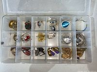 Plastic organizer box with 20 compartments holding assorted small jewelry pieces including earrings, butterfly brooch, heart-shaped pendants, and an engraved money clip. Varied colors and metal tones visible.