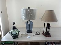 Wide shot of three nautical-themed table lamps on a white table: blue glass lamp with white shade wrapped in plastic, black metal lamp with rounded glass body, and copper oil lantern style lamp with linen shade