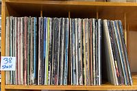 Full view of 38 vinyl albums stored vertically on a wooden shelf, showing varying spine colors and patinas
