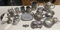 Wide shot showing entire collection of vintage silver plated and pewter items on table, including platter, teapot, butter dish, mugs and bowls