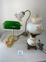 Front view of three lamps: green glass shade brass banker lamp, brass floor/desk lamp with clear glass bird shade, and floral painted glass globe bedside lamp on brass base.