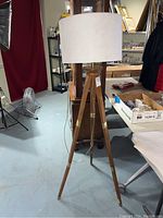 Full view of the triple leg floor lamp showing wooden legs with brass hardware and white drum lampshade in a room with other items.