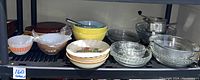 Wide view of multiple mixing bowls, baking dishes, pie plates including clear glass and colored ceramic bowls on black shelving unit