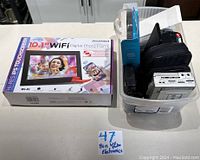 Photo of a box of Sylvania 10.1 WiFi digital photo frame and assorted electronics in plastic bin including cassette players, remote controls, and small devices.