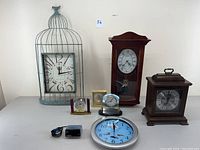 Photo showing all seven clocks in the lot arranged on a white table against a white wall, including a birdcage-style clock, wooden wall clocks, mantel clock, plug-in digital clock, and three small desk clocks.