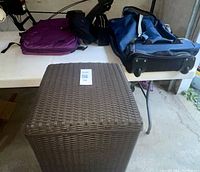 Overview of brown wicker laundry basket and three bags on table: purple backpack, black bag insert, blue pullover technology bag with wheels.