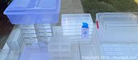 Wide view of multiple plastic storage units arranged on a table outdoors, showing stackable containers, drawer organizers, and compartment trays largely clear or translucent purple.